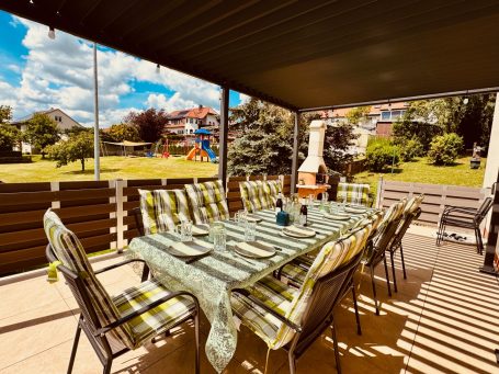 Große, überdachte Terrasse mit Grill und großem Tisch – ideal für gemeinsame Abende. Blick auf den Spielplatz im Garten, Kinder können spiel